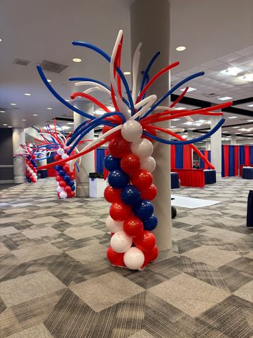 Red, white, and blue balloon columns with fountain tops line an indoor event space.