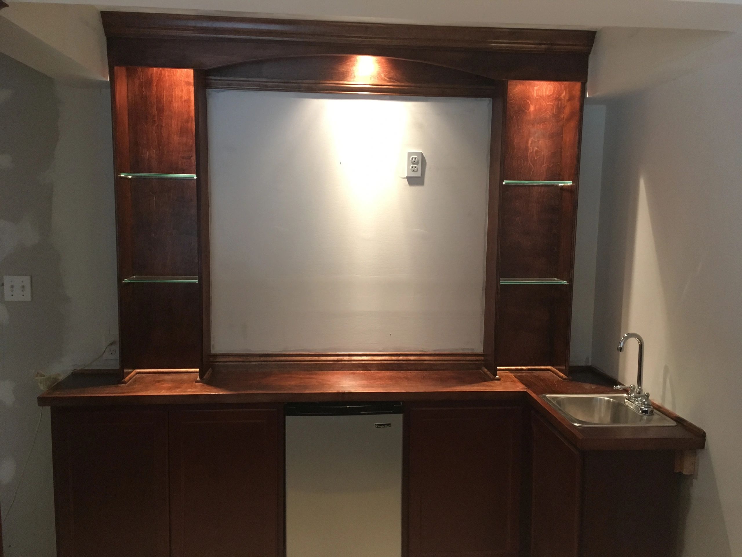 Wooden bar setup with shelves, mini fridge, and sink under warm lighting.
