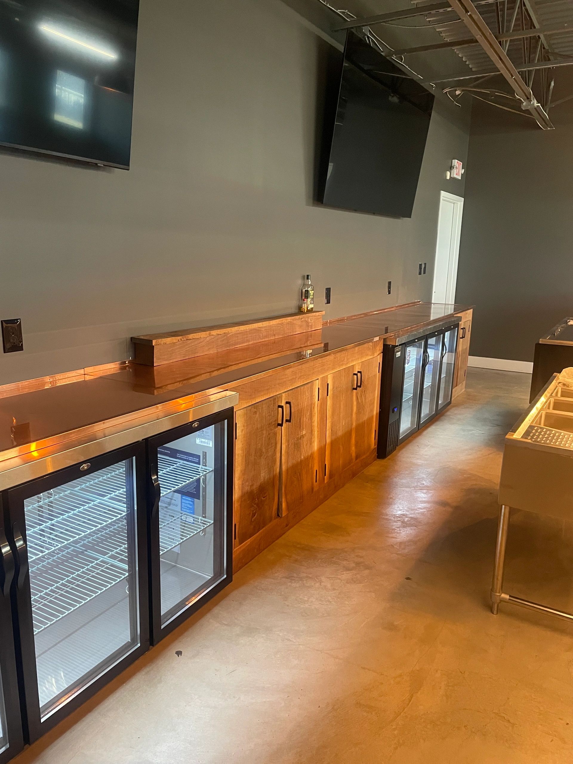 Empty bar counter with wooden cabinets and glass-door refrigerators beneath two wall-mounted TVs.
