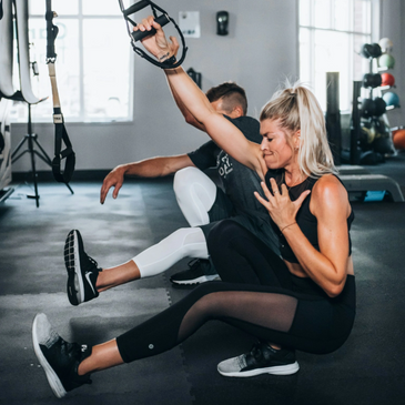 a woman and man performing pistol squats using a suspension trainer in a gym