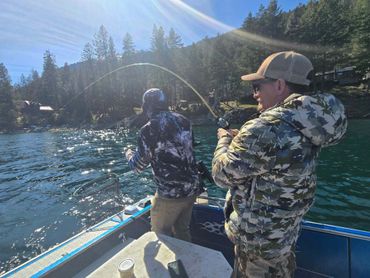 Two men fishing on a boat in a sunlit lake surrounded by trees.