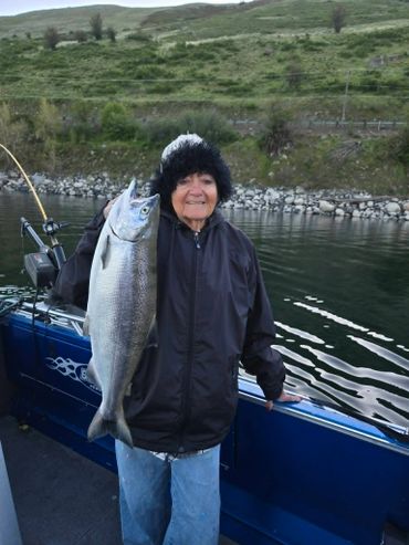 Elderly woman proudly holds a large fish on a boat in a scenic river setting.