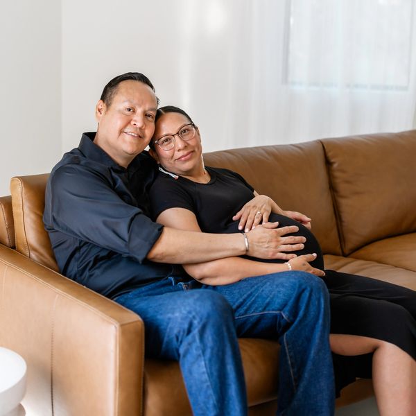A husband and wife sitting on a couch cradling a baby bump. 