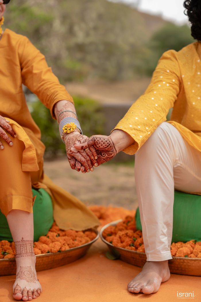 Couple holding hands adorned with intricate henna designs, dressed in traditional yellow attire.