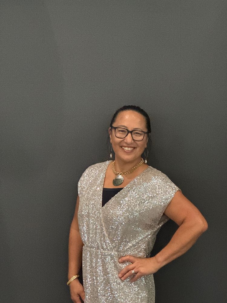 woman smiling in gold dress, gold and large oyster shell necklace, hair slicked back 

