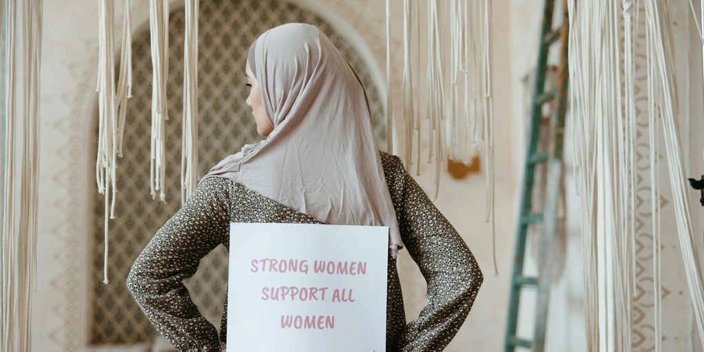 Woman wearing hijab holds sign reading 'Strong women support all women.'