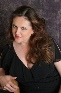 Woman with wavy brown hair in a black dress posing against a textured backdrop.