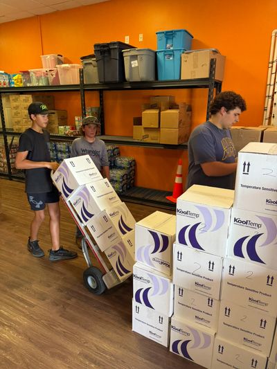 Three young people organizing and moving stacked boxes in a storage room with orange walls.
