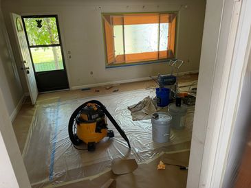 Room under renovation with vacuum and painting supplies on plastic-covered floor.
