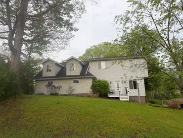 Two-story house with beige siding surrounded by trees and lawn.