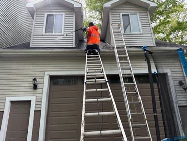 Worker in orange jacket on ladder fixing house siding near windows.