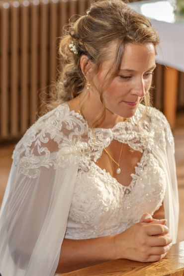 A bride in a lace wedding dress and cape, sitting with hands clasped.