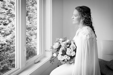 Bride in a lace gown gazes out a window holding a flower bouquet.