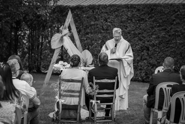 Couple seated outdoors during a wedding ceremony with a priest speaking.