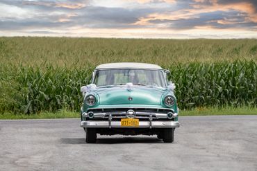 A vintage green car parked on a rural road with a cornfield background.