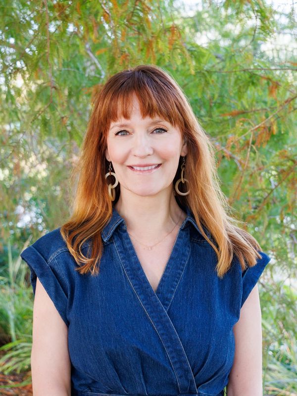 Smiling woman with red hair in a denim dress stands outdoors with greenery background.