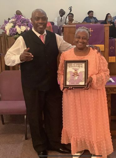 A man and woman smiling, woman holding a framed certificate in a church setting.