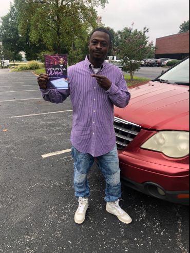 Man in purple shirt holding a book, standing next to a red car.