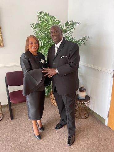 A well-dressed couple smiling, standing indoors near a plant.