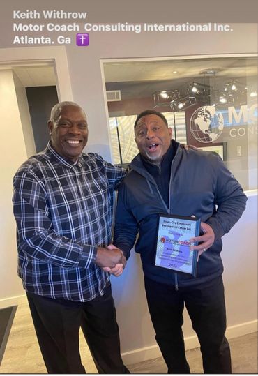 Two men shaking hands in an office, one holding an award plaque and smiling.