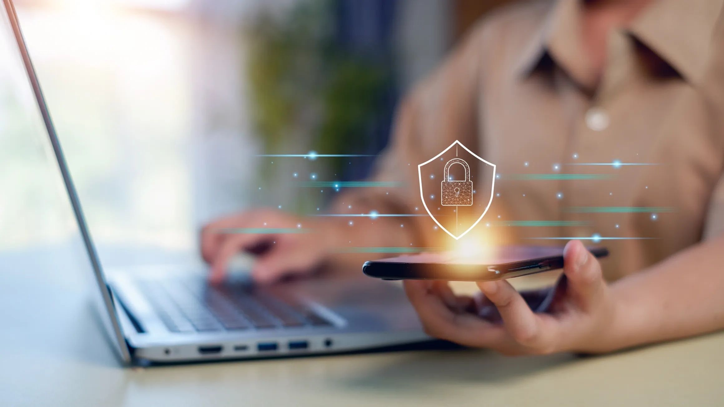 A person using a laptop & smartphone with a glowing digital padlock icon representing cybersecurity
