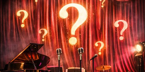 Stage set with vintage microphones, piano, and question marks on curtain.