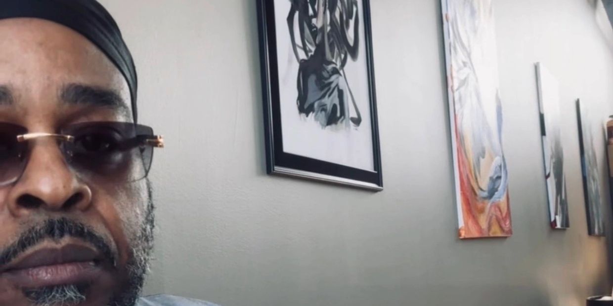 Man wearing sunglasses and a headwrap by a wall with abstract paintings.