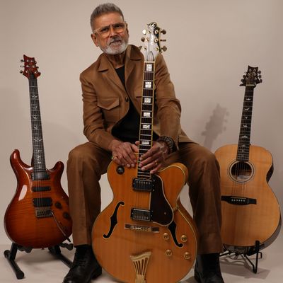 Man in brown suit posing with three guitars against a plain background.