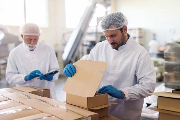 Two workers in lab coats and hairnets pack boxes in a warehouse.