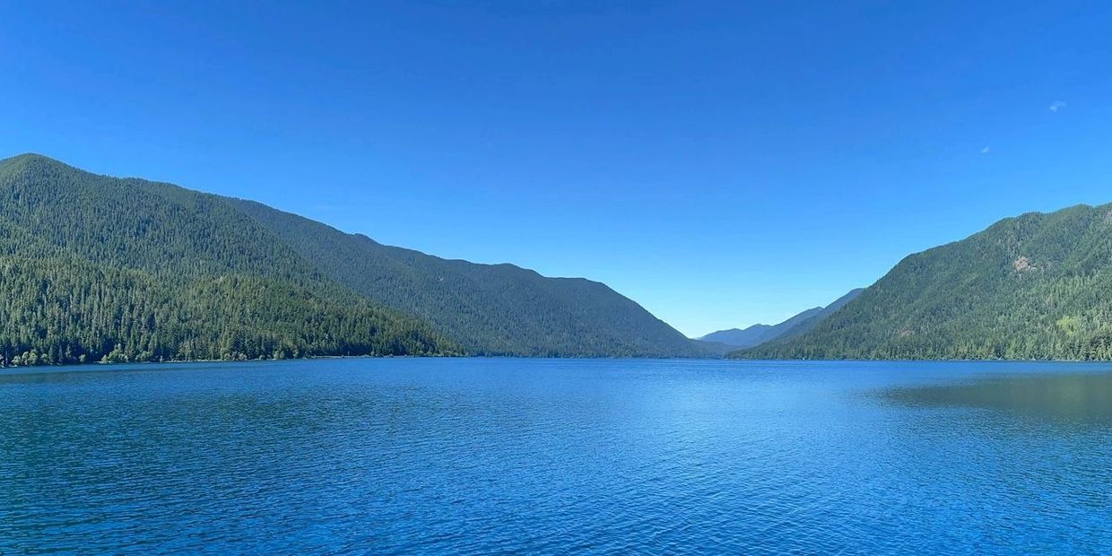 Green mountains dipping to a valley at the far end of a blue lake with a blue sky.