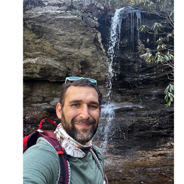 Man smiling with a small waterfall in the background during a hike.