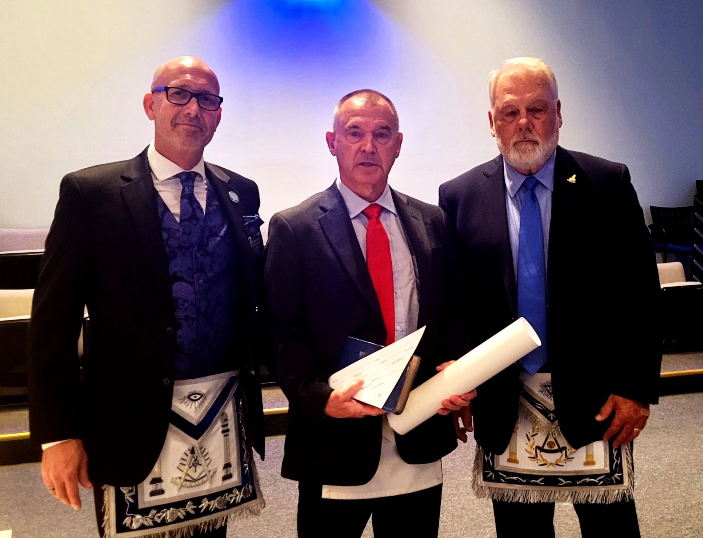 Three men in formal attire with Masonic aprons holding certificates and scrolls.