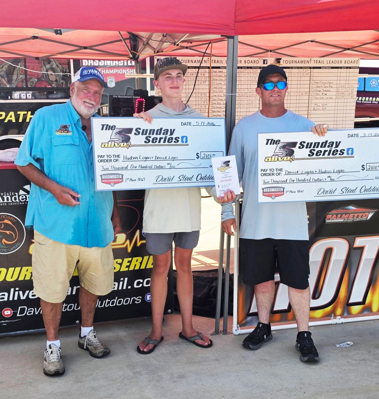 Three men holding large checks and a trophy under a tent.
