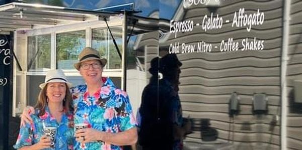 Couple in matching floral shirts stands by a coffee truck on a sunny day.