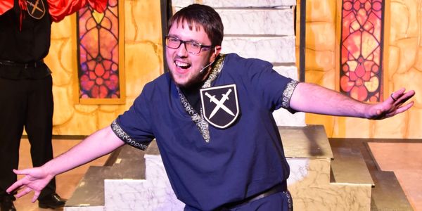 A man in medieval costume performing on stage with arms outstretched.
