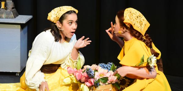 Two women in yellow costumes perform a lively scene with flowers on stage.