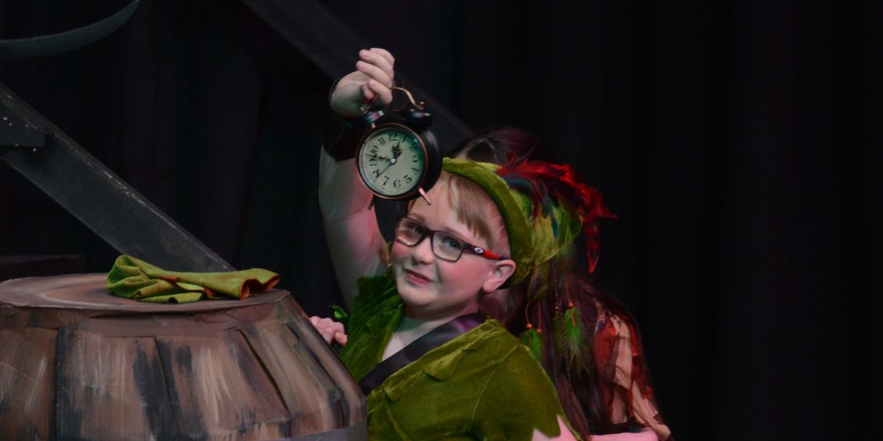 Child in green costume holding an alarm clock during a play.