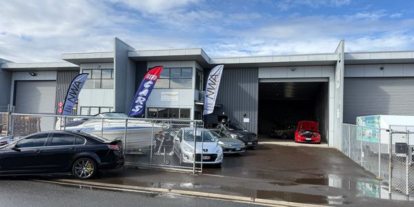 Used car dealership with several cars and a boat outside a warehouse building.