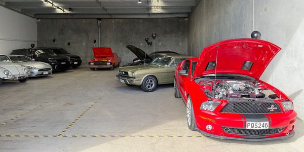 A lineup of classic and modern cars in a spacious garage with some hoods open.