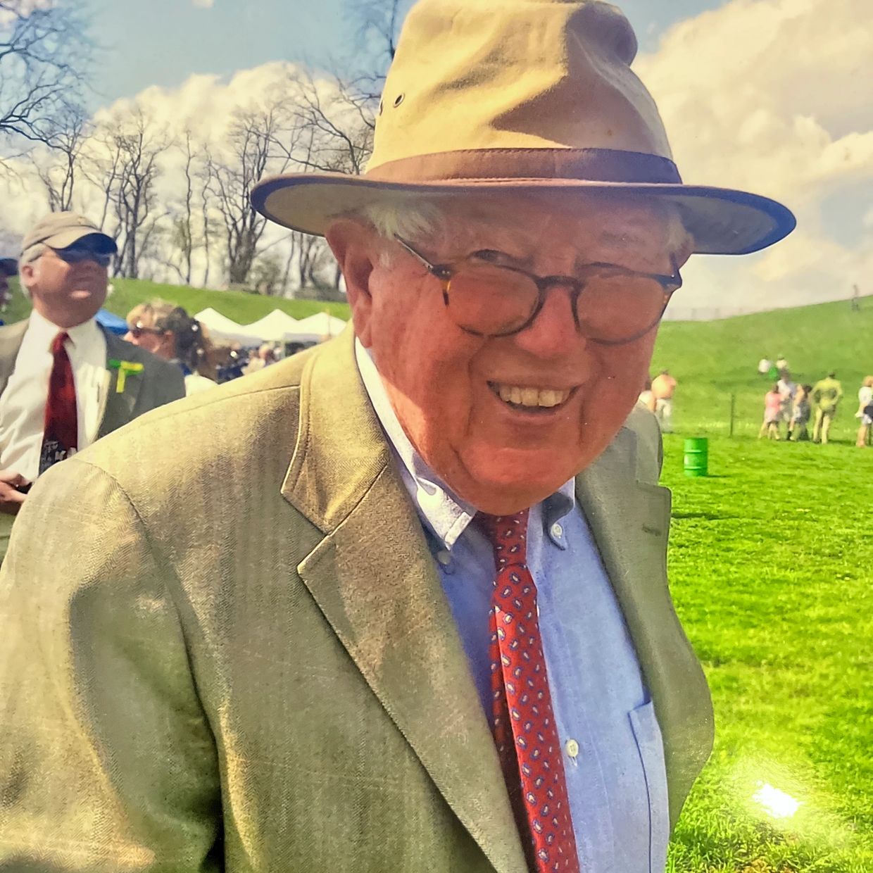 Smiling elderly man in a hat and suit at an outdoor event.