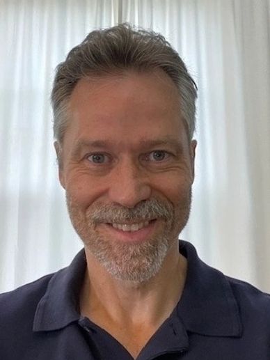Smiling man with gray hair and beard in front of white curtains.