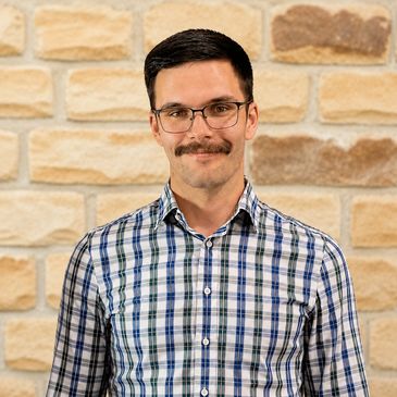 Man with glasses and mustache smiling against a brick wall.