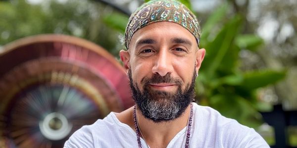 Bearded man in white shirt and headscarf, wearing spiritual necklaces, smiling outdoors.