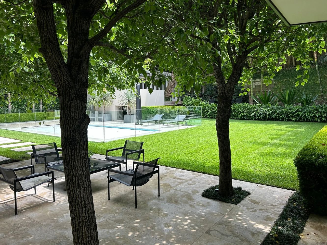 A well-maintained backyard with green lawn, pool, and patio furniture under shaded trees.