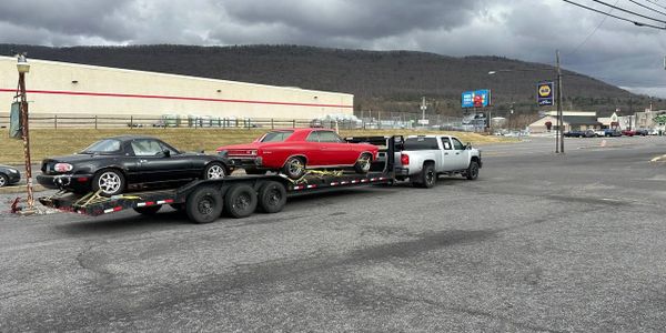 Two cars, one black and one red, loaded on a trailer hitched to a silver truck under a cloudy sky.