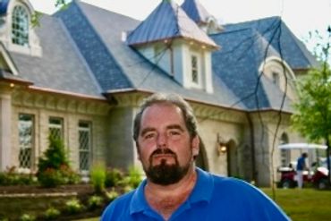 Man with beard standing outside a large house on a sunny day.