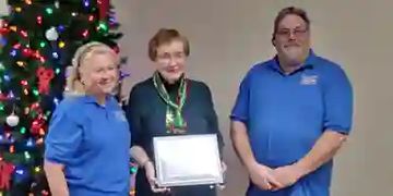 Three people standing indoors with a Christmas tree, one holding a certificate.