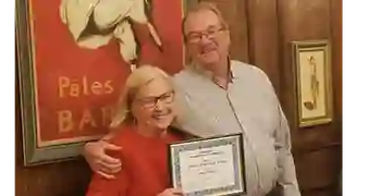 Smiling elderly couple holding a certificate in a warmly decorated room.
