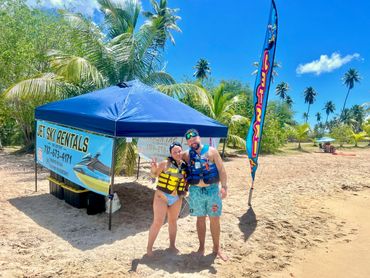 Couple posing on beach near jet ski rentals tent under clear blue sky.