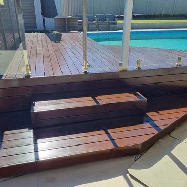 Wooden poolside deck with glass railing and steps.